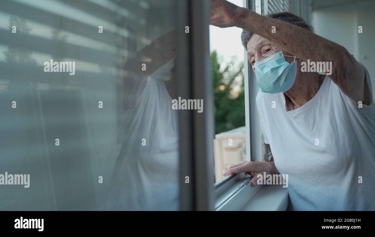 Happy senior woman in mask waves to family from window hospital. Masked elderly female glad that children came visit her in nursing home. Grandmother Stock Photo