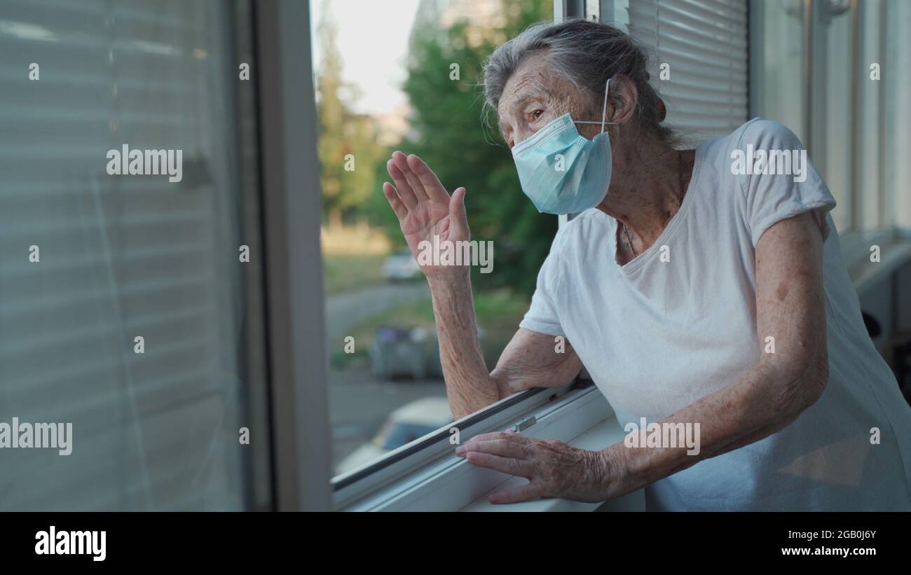 Lonely senior woman in mask stands by window and waves her hand to family who came visit her at hospital. 90 year old female in disguise says goodbye Stock Photo