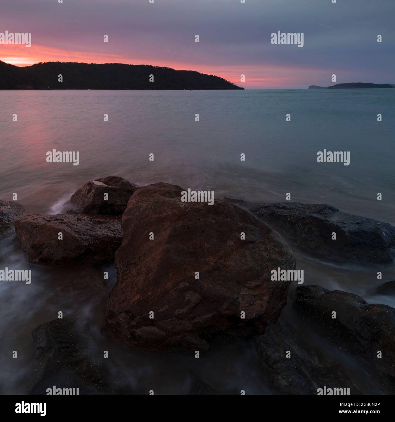 pink glow in the sky at sunrise on the beach with rocks in foreground Stock Photo