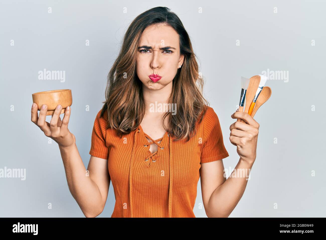 Young hispanic girl holding make up products puffing cheeks with funny face. mouth inflated with air, catching air. Stock Photo