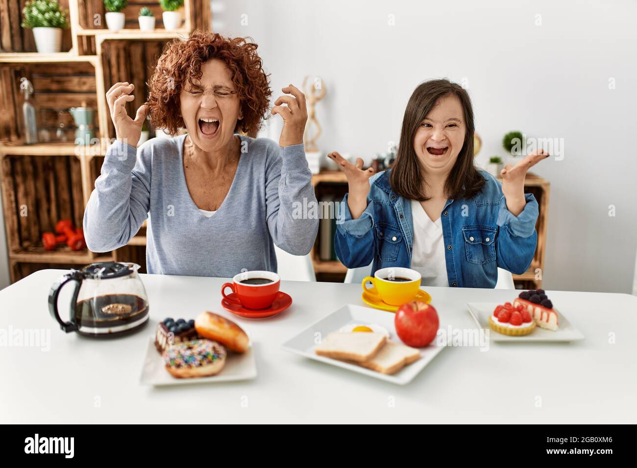 Family of mother and down syndrome daughter sitting at home eating breakfast celebrating mad and crazy for success with arms raised and closed eyes sc Stock Photo