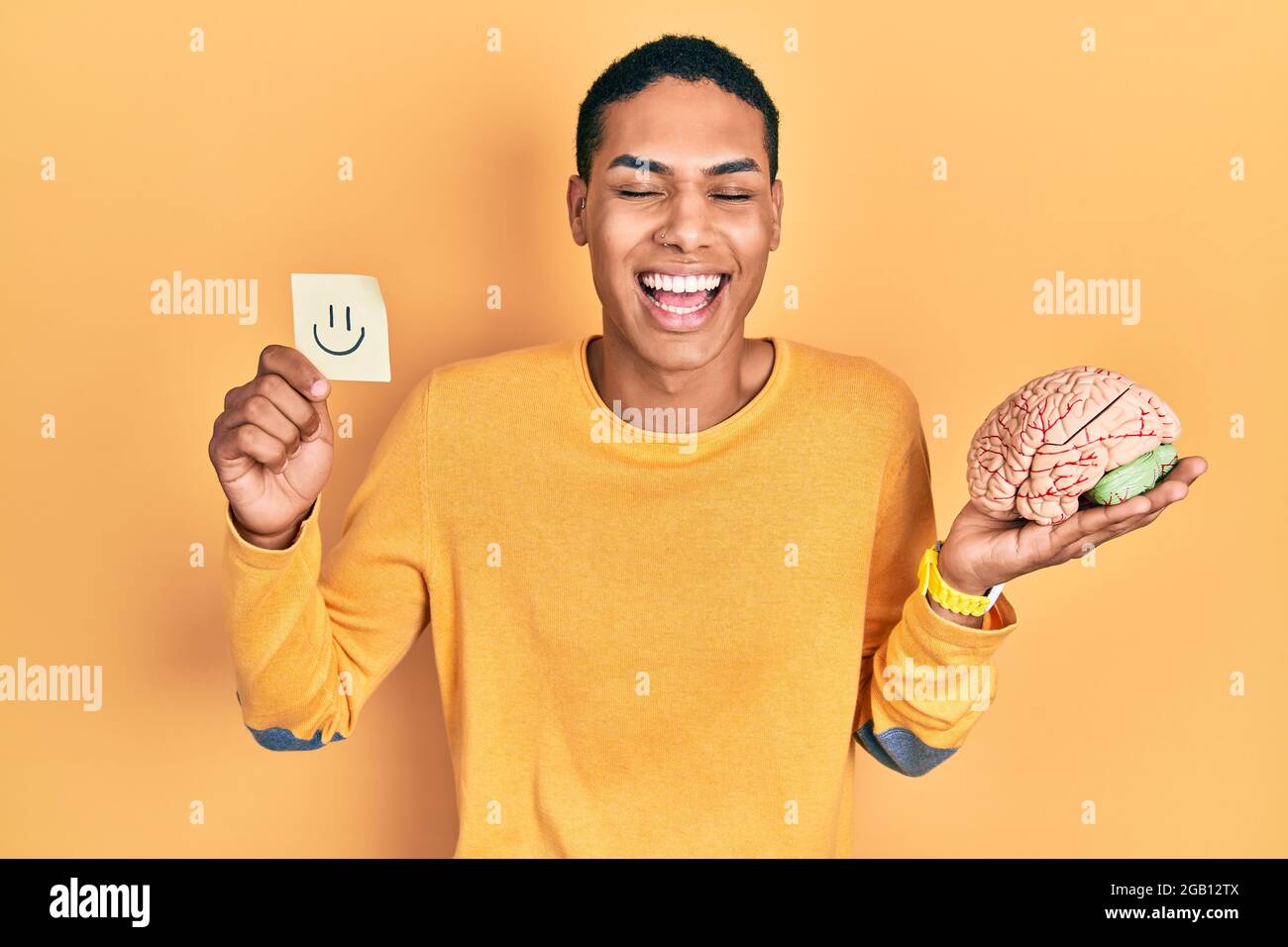 Young african american guy holding smile reminder and brain smiling and laughing hard out loud because funny crazy joke. Stock Photo