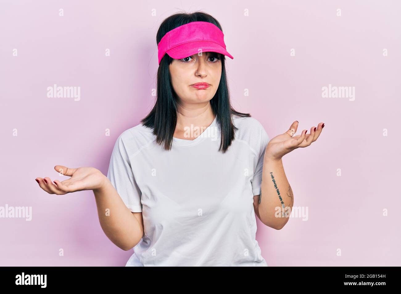 Young hispanic woman wearing sportswear and sun visor cap clueless and confused expression with arms and hands raised. doubt concept. Stock Photo