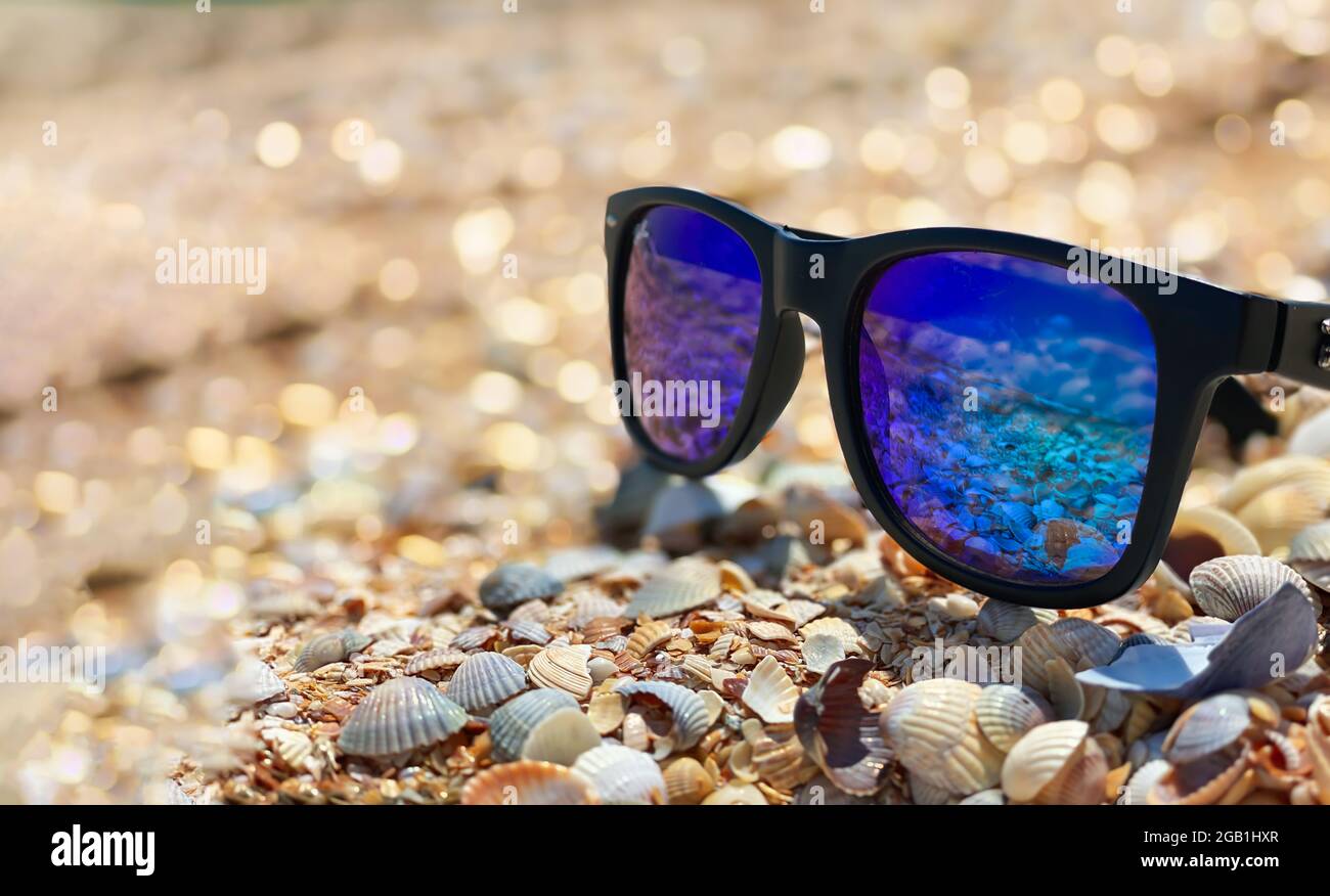 Sunglasses with blue lenses lie on a sandy shell beach on the sea front in sunny weather. Stock Photo