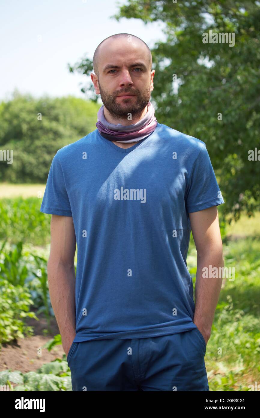 White man in a blue T-shirt and shorts. A bald man with stubble on his face holds his hands in his pockets. Looking into the camera. Stock Photo
