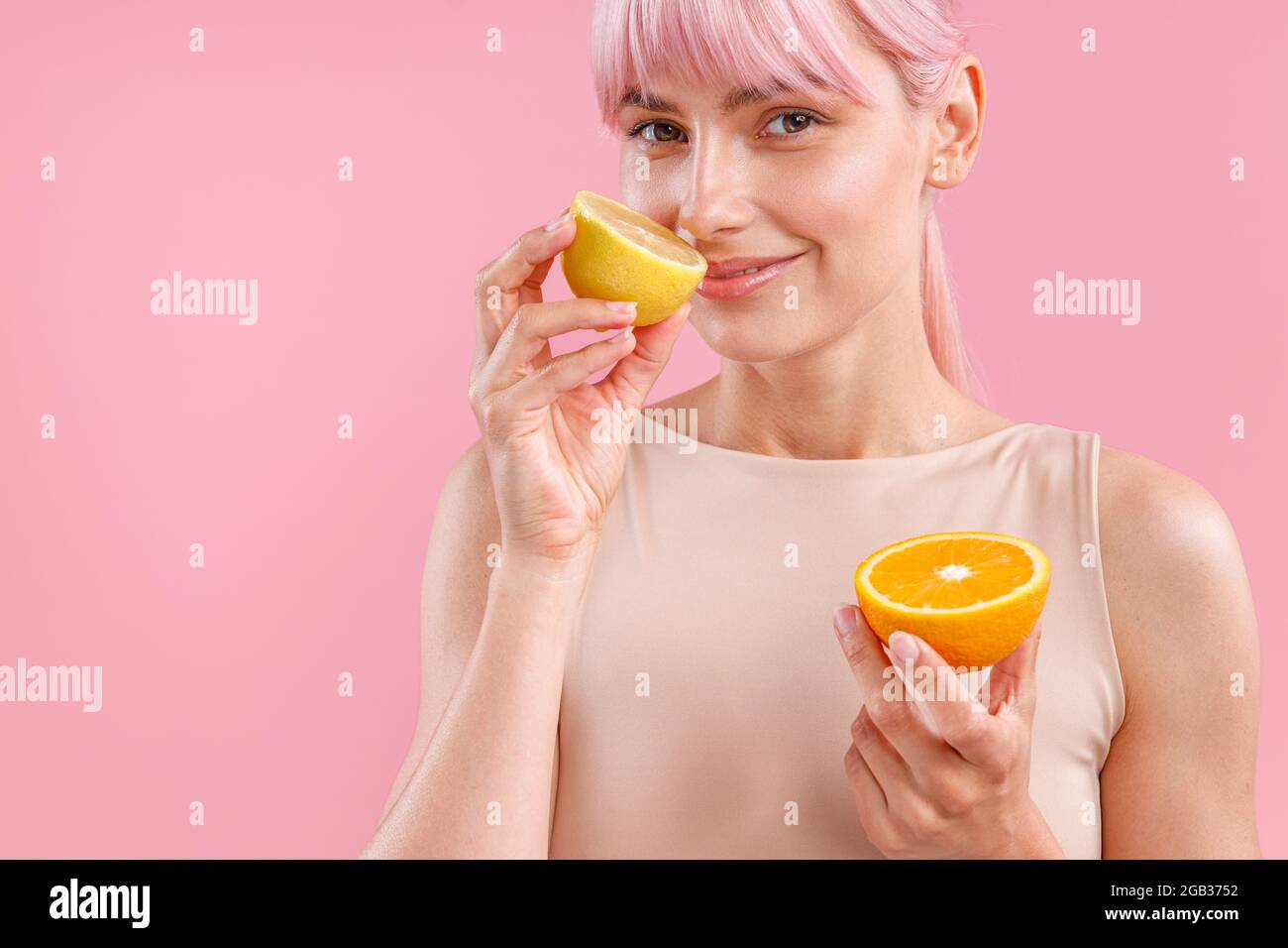Portrait of smiling woman with pink hair holding half of ripe orange and smelling fresh lemon, posing isolated over pink background Stock Photo