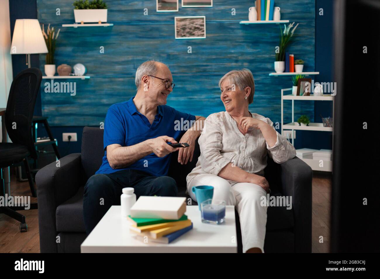 Old couple enjoying retirement at home sitting on sofa watching television. Senior people having fun together with tv movie technology for relaxation entertainment being indoors Stock Photo