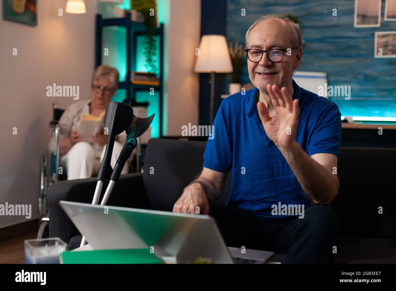 Retired old couple with handicap sitting indoors having walking support supplies. Senior man with crutches using laptop computer video call technology and elder woman disabled in wheelchair Stock Photo