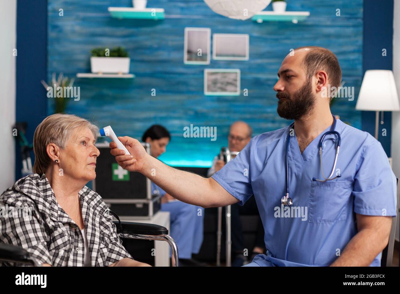 Assistant man helper checking temperature using medical infrared thermometer discussing with senior woman during therapy. Social services nursing elderly retired female. Healthcare assistance Stock Photo