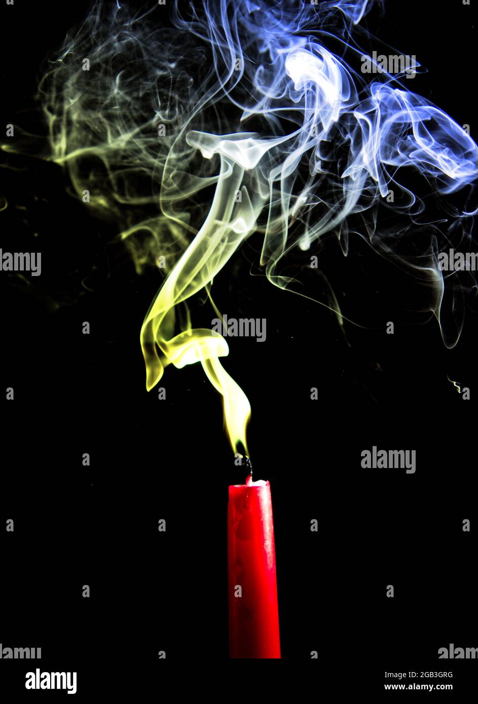 Blue and yellow smoke rise from a red candle that has been blown out.  Black background Stock Photo