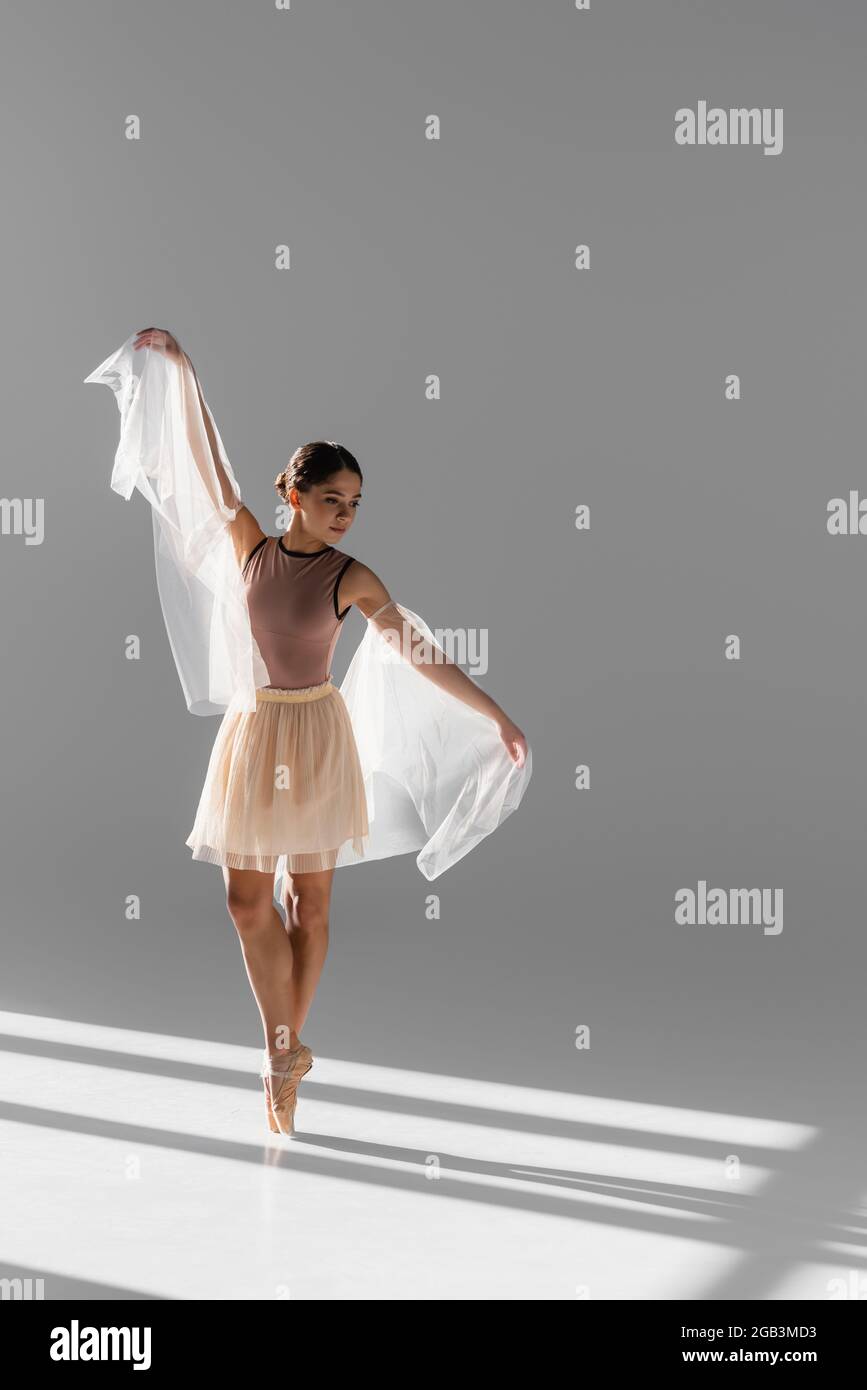 Pretty ballerina in beige skirt dancing with cloth on grey background