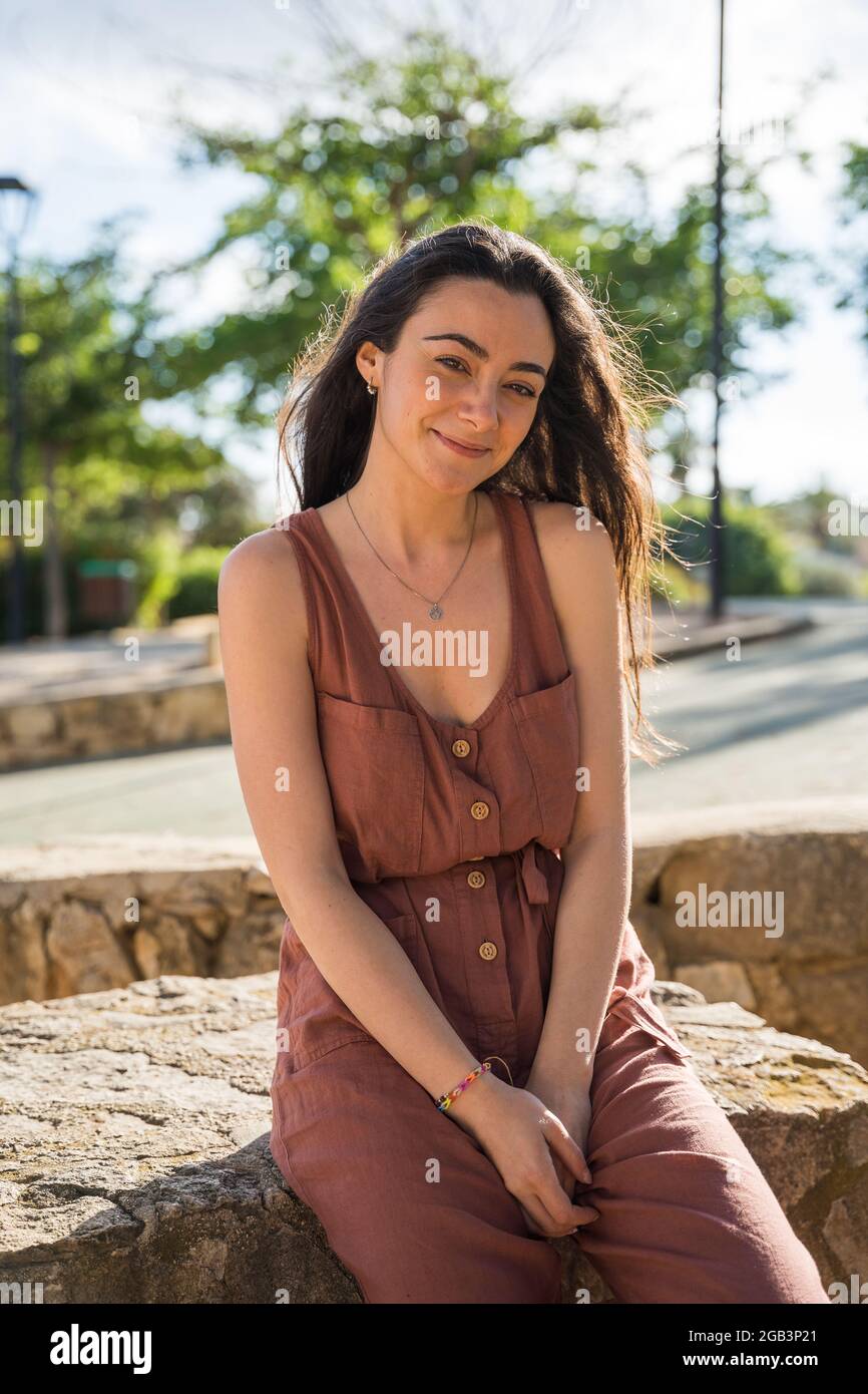 Young woman sitting looking at camera smiling. Caucasian with dark hair. Wearing a red dress. She is sitting on a stone bench in a park. Javea, Alican Stock Photo