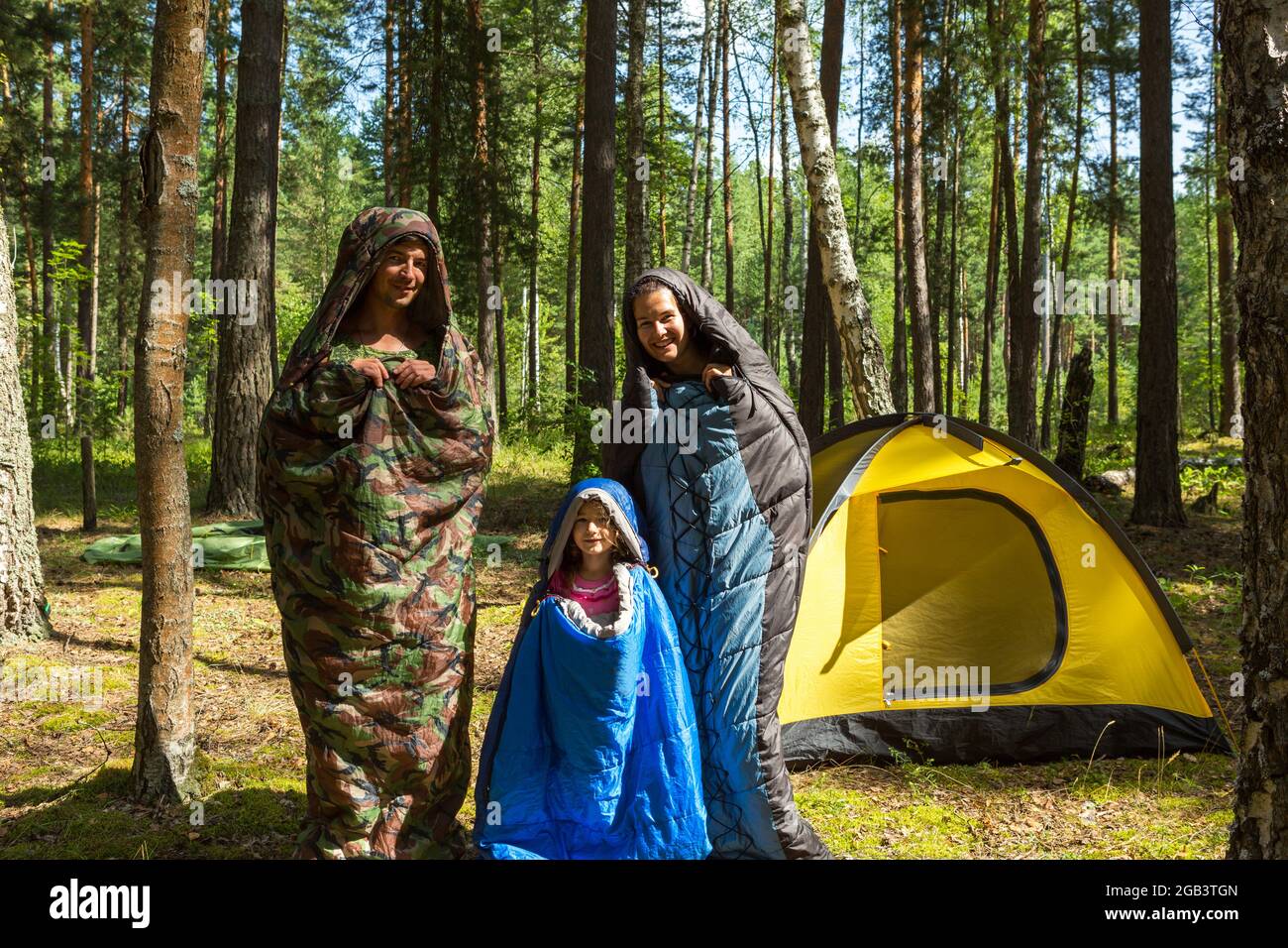 Family of tourists from a father, mother and little daughter pose in sleeping bags near a tent. Family outdoor recreation, domestic tourism, camping, Stock Photo