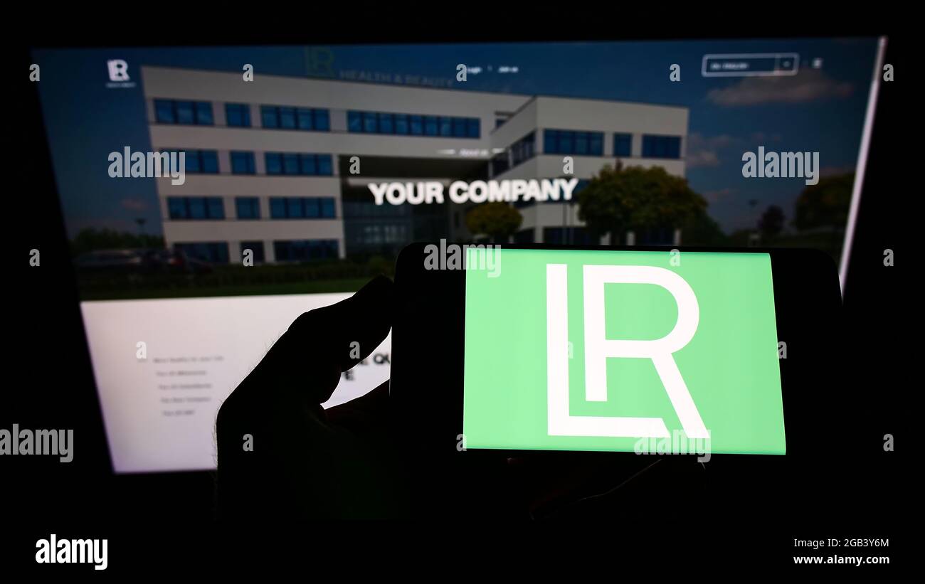 Person holding smartphone with logo of cosmetics company LR Health and Beauty Systems GmbH on screen in front of website. Focus on phone display. Stock Photo