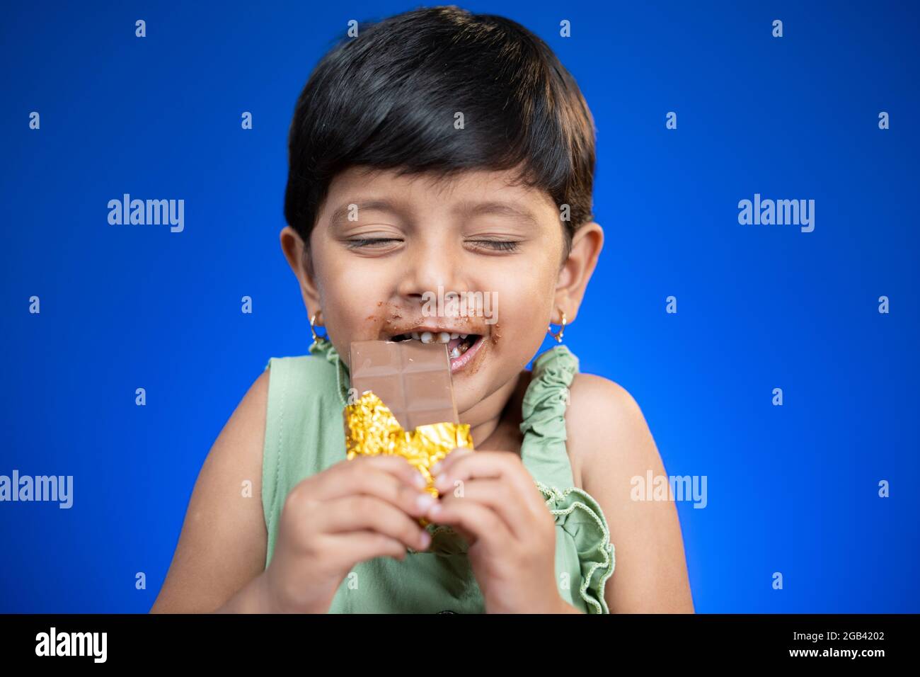 Happy girl kid enjoying chocolate bite by closing eyes on blue background - concepts of children's love and addictions on chocolates Stock Photo