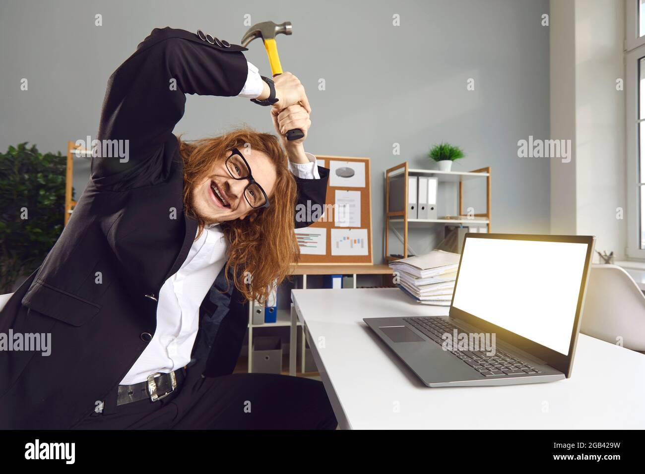 Portrait of a funny crazy office worker sitting at a desk breaking a laptop with a hammer. Stock Photo