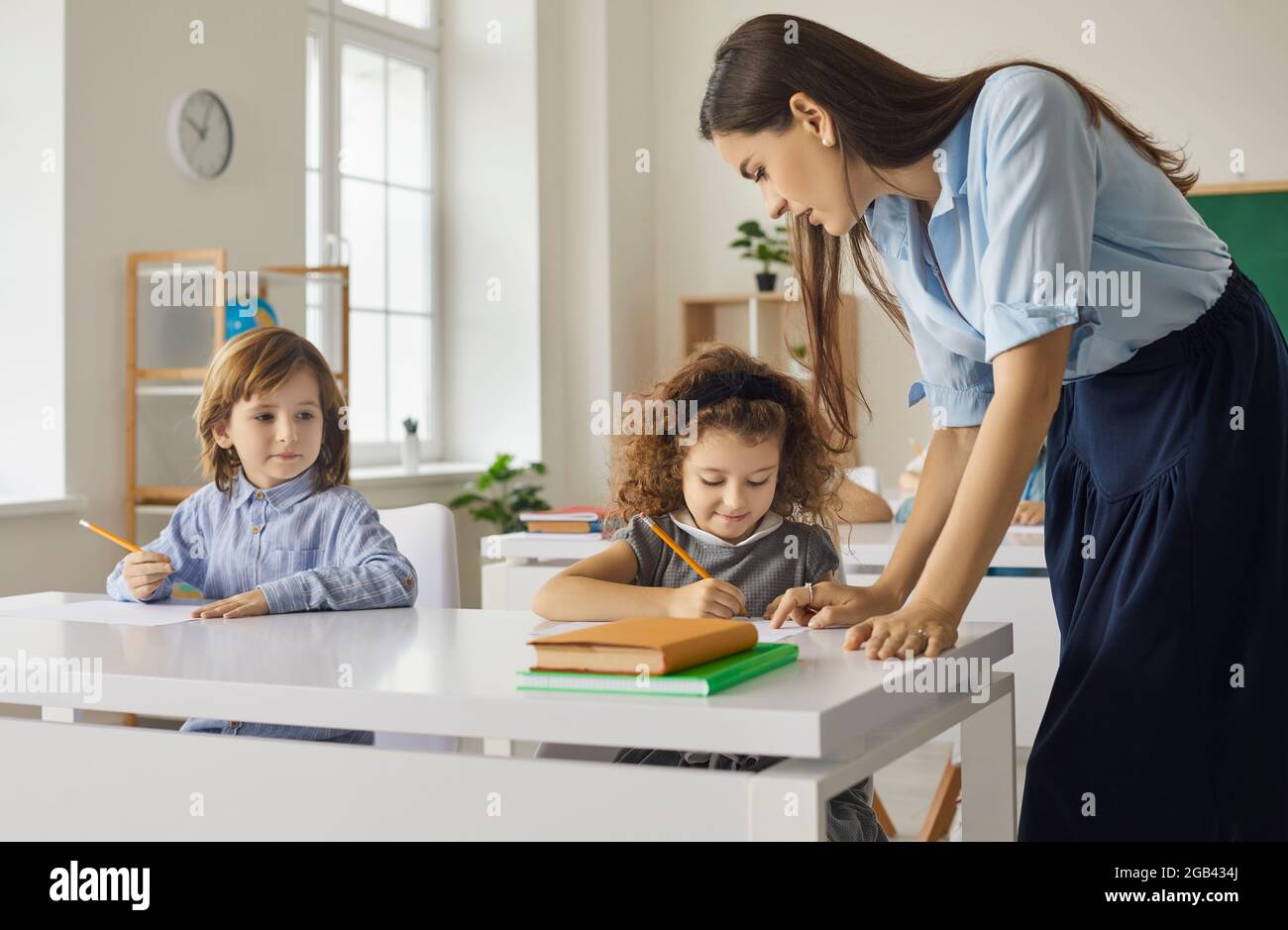 Elementary school students sit at desks and write or draw under the supervision of a school teacher. Stock Photo