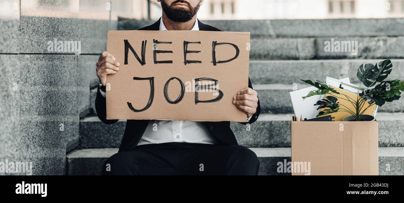 Unemployment concept. Mature businessman sitting on stairs with poster Need Job and box of personal belongings, crop Stock Photo