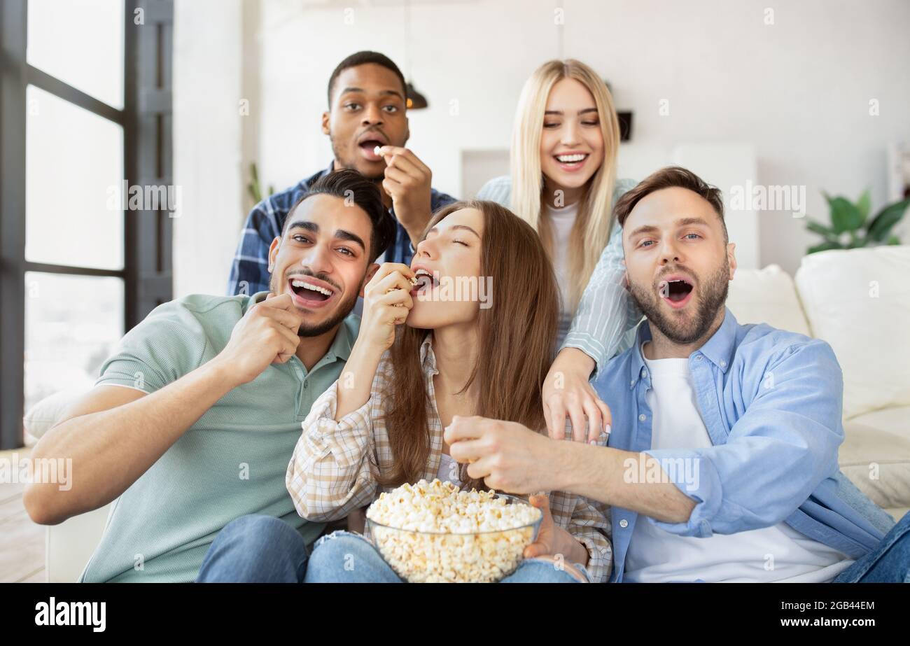 Diverse young friends eating popcorn while watching TV, having fun together at home Stock Photo