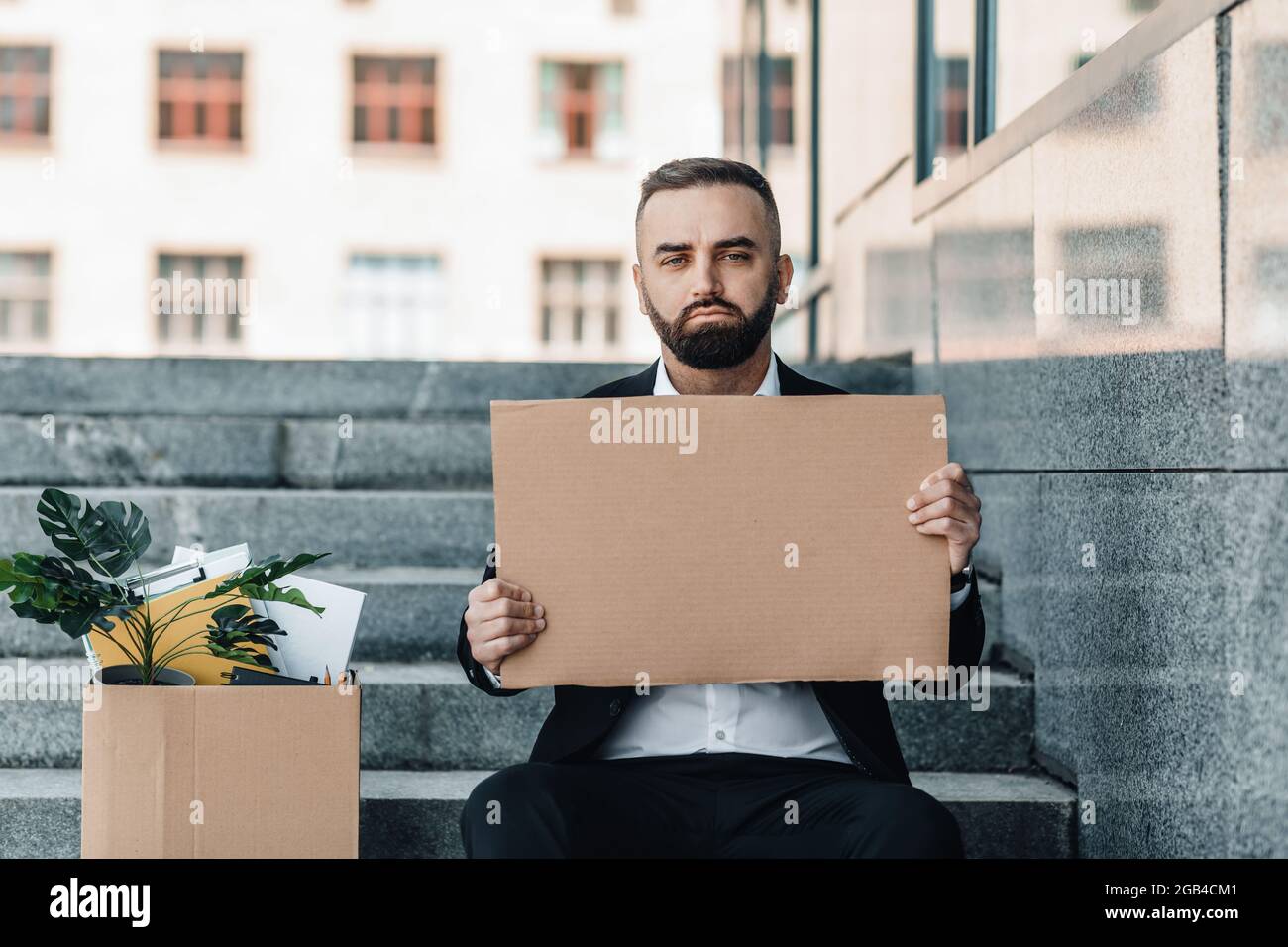 Sad middle-aged businessman sitting with empty poster and box of personal belongings on stairs near office center Stock Photo