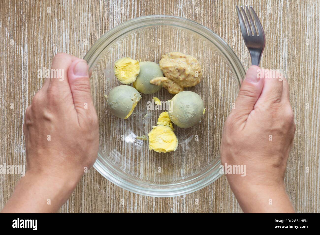 Top view of woman hands holding fork to crush boiled yolks and mustard in glass bowl to make flavoring for okroshka Stock Photo