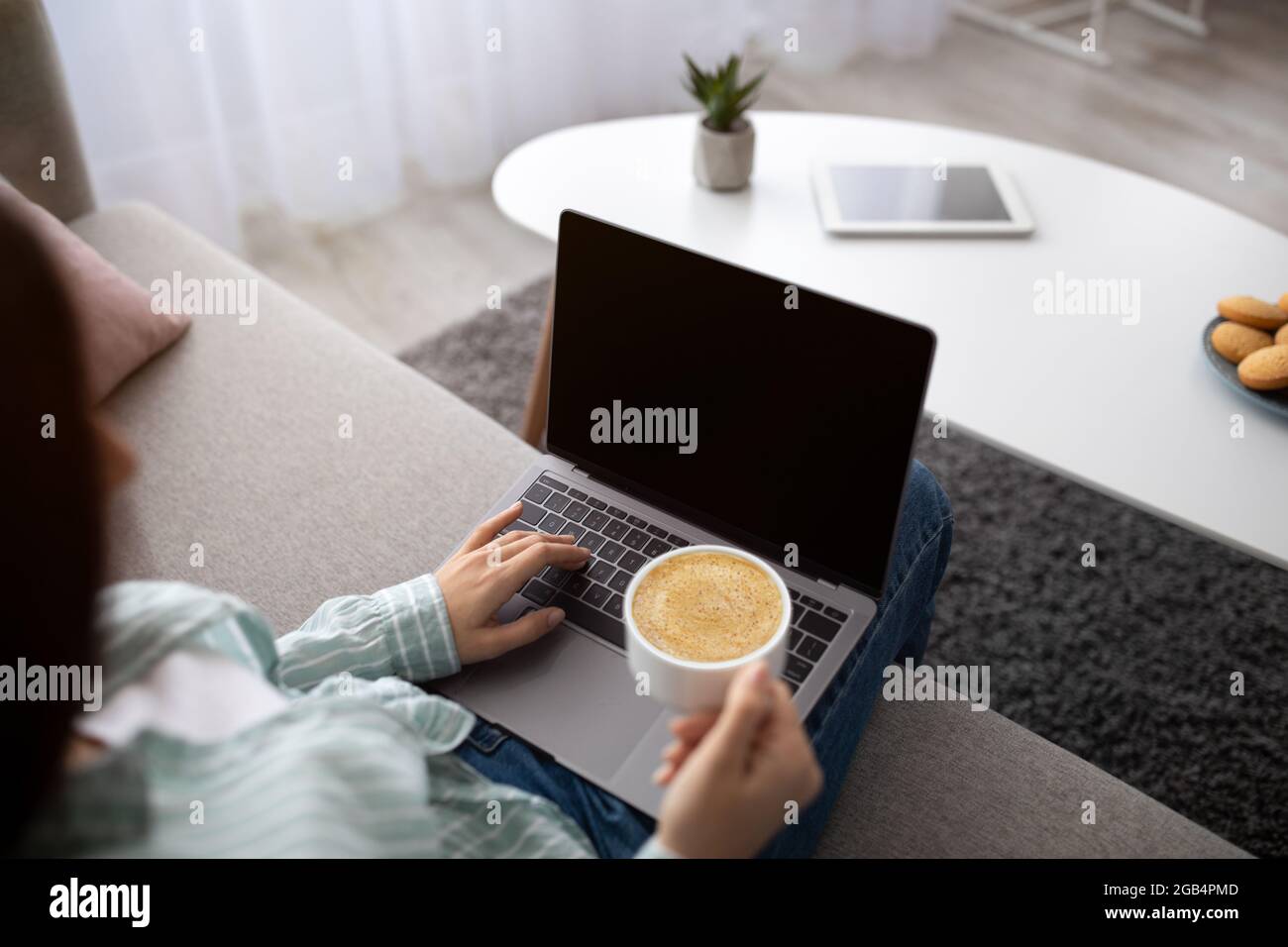 Young Indian woman with coffee sitting on sofa and using laptop computer with blank screen at home, mockup Stock Photo