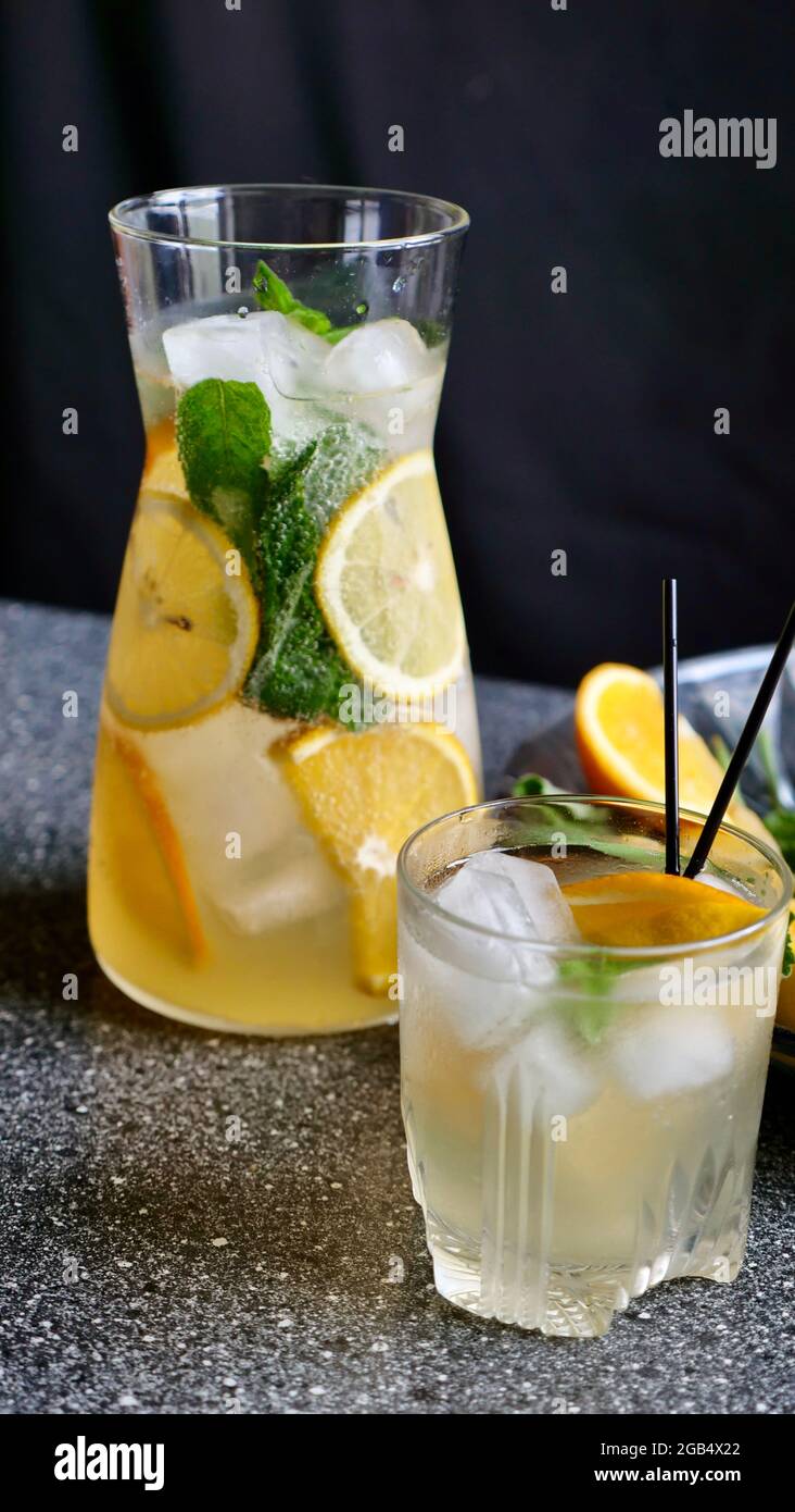 Lemon lemonade with mint, lemon and ice in a decanter and a glass with black plastic tubes on a background of fruit. Cooling, refreshing, fruit, summer drink. Vertical close up Stock Photo