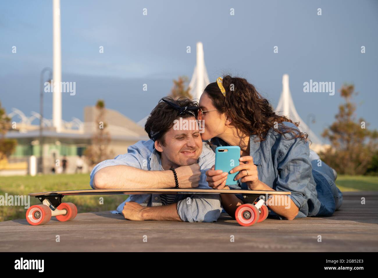 Stylish young couple in love lying on skateboard outdoors looking at smartphone chilling at sunset Stock Photo