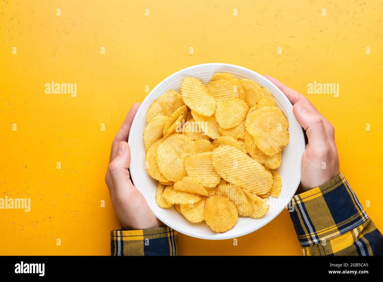 Potato chips or crisps in bowl in male hands over yellow background, top view, copy space. Superbowl snack, junk food Stock Photo