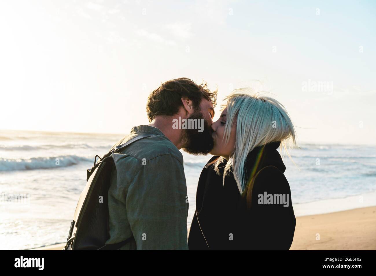 Romantic happy couple in love kissing on shoreline at the wild beach - Boyfriend and girlfriend wearing cool clothing and vintage backpack standing in Stock Photo