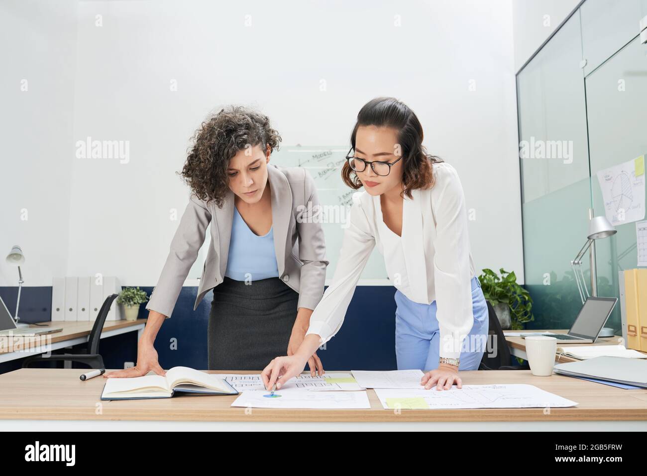Serious businesswomen bending over table with charts and reports and analyzing financial data Stock Photo
