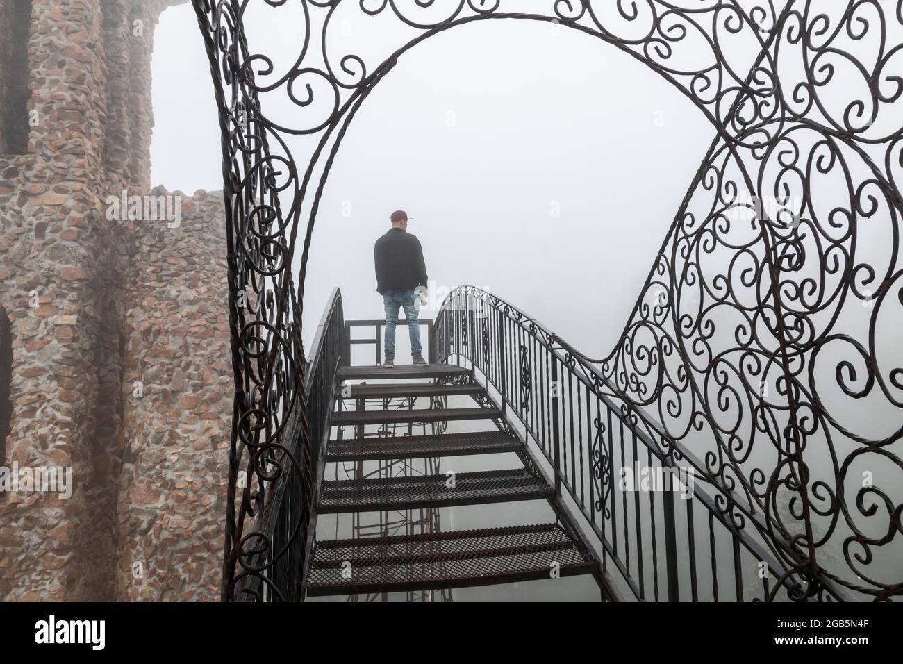 Rye, Colorado - A main stands on an open stairway, high above the ground, at Bishop Castle on a foggy morning. The Castle is an elaborate stone and me Stock Photo