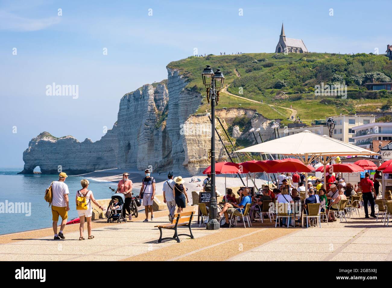 People having a drink at a beach cafe on the seafront in Etretat, France, with Notre-Dame de la Garde chapel overlooking the Amont cliff and its arch. Stock Photo