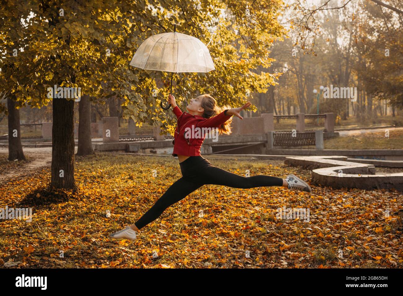 Hello fall, autumn season, fall mood, positive emotions. Teenager girl jumping with transparent umbrella and enjoying life in autumn sunny park Stock Photo