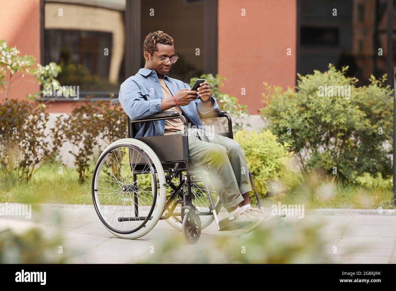 Content handicapped African-American man sitting in wheelchair on street and reading message on phone while chatting online Stock Photo