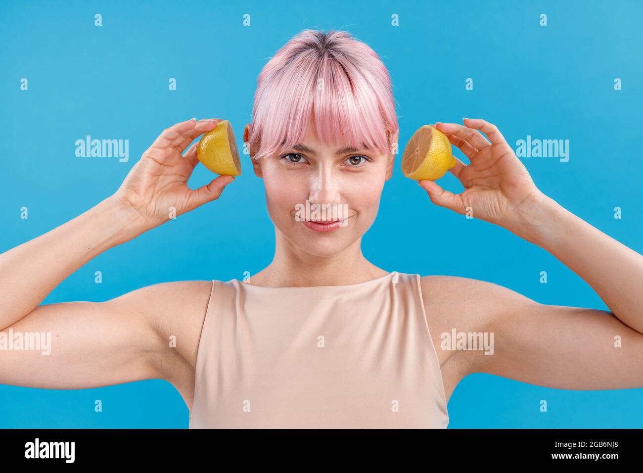 Portrait of young woman with pink hair smiling and holding halves of fresh lemon near her ears, posing isolated over blue background Stock Photo