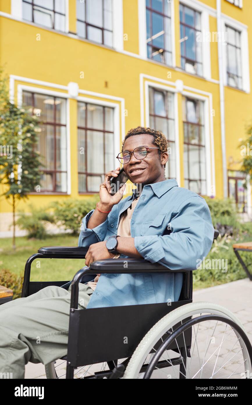 Portrait of positive handsome young disabled African-American man in glasses sitting in wheelchair and calling by phone Stock Photo