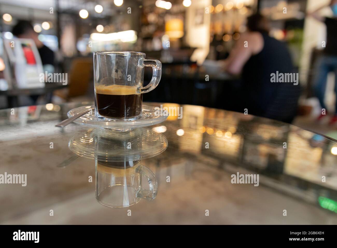 Close up landscape image of a glass cup of espresso with natural foam sitting on top of a mirror table in a posh cafe. Stock Photo