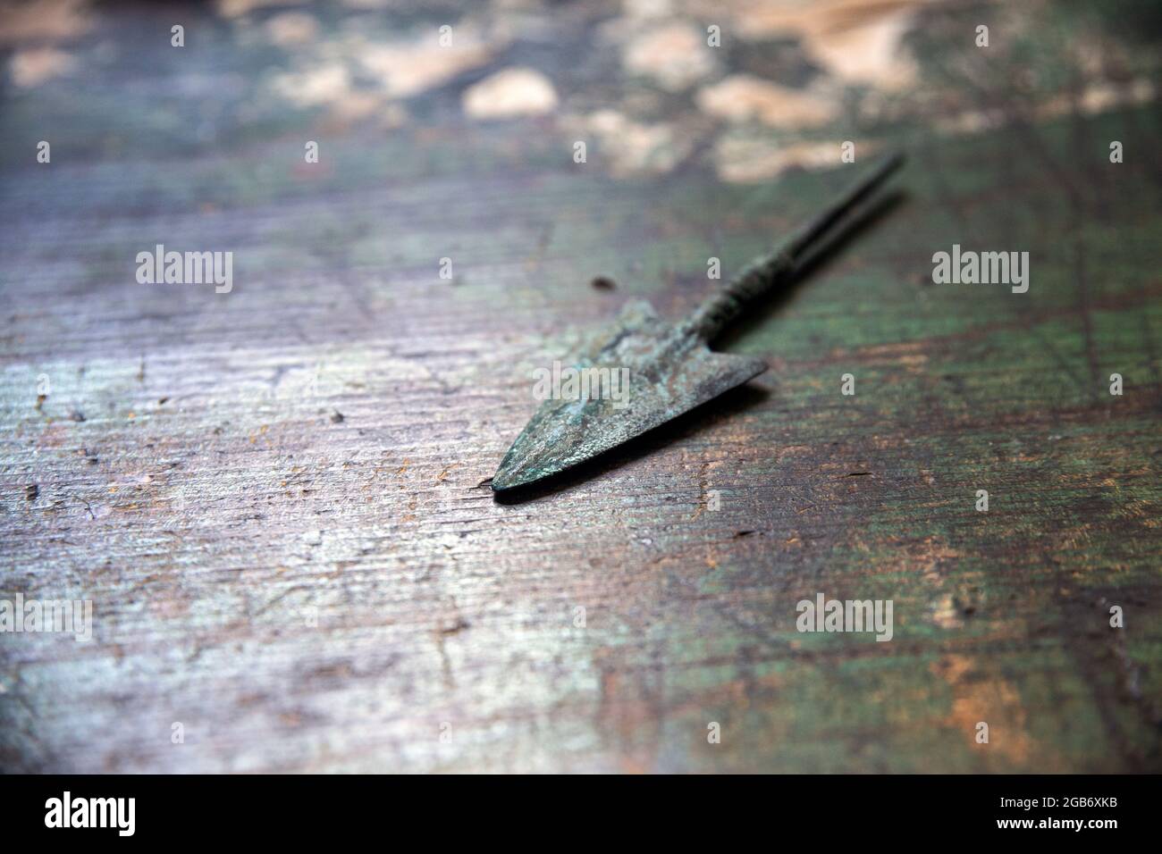 Close up of an arrowhead from antiquity, made of bronze, oxidized color on similar color wooden surface with fainted green paint Stock Photo