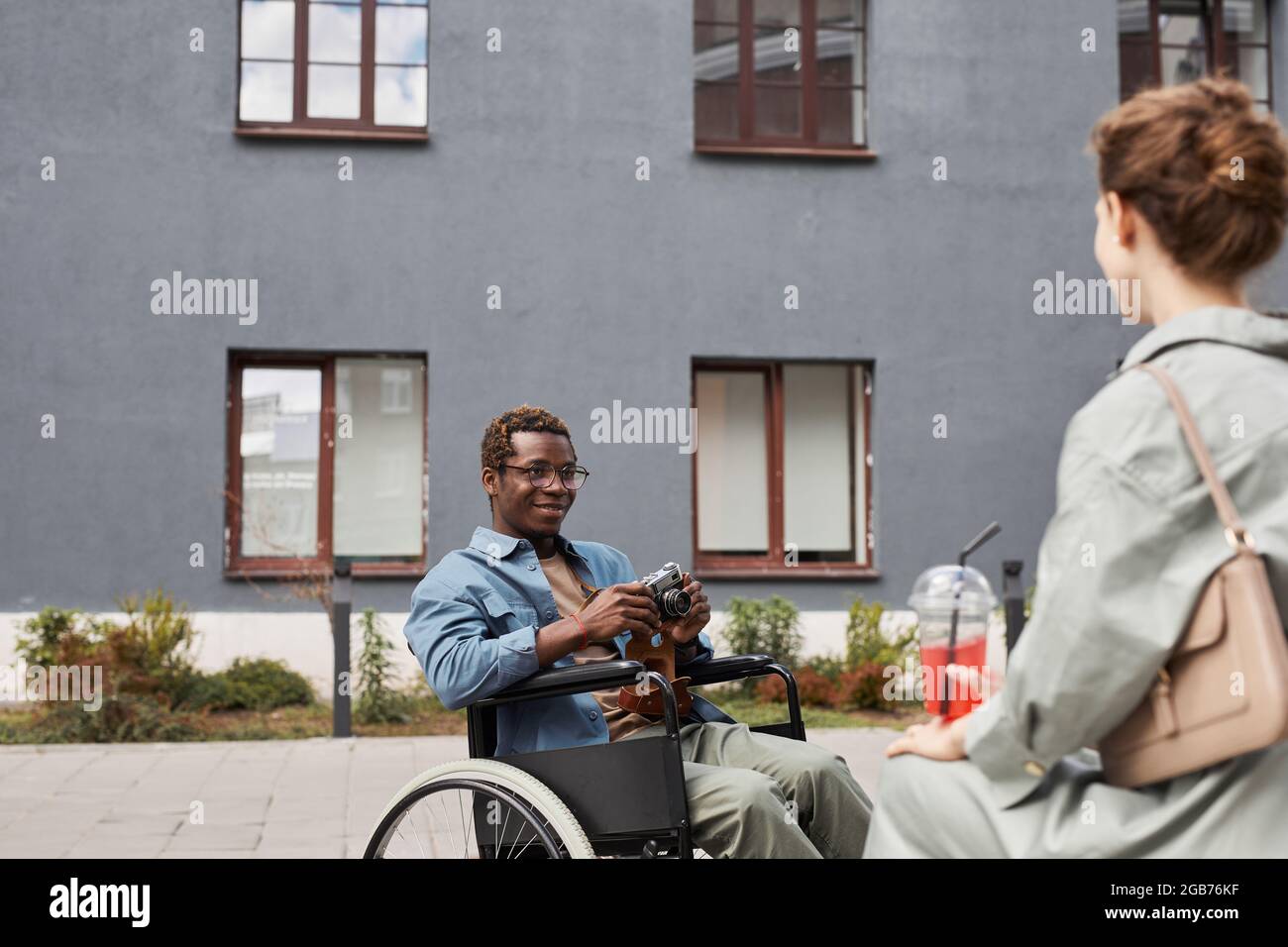 Smiling young handicapped African-American man in glasses sitting in wheelchair and photographing woman outdoors Stock Photo