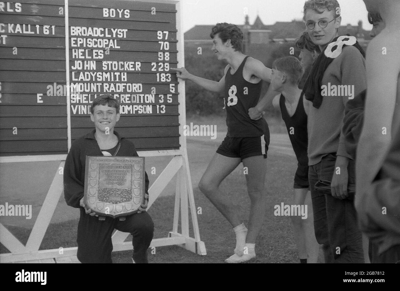 1964, in his cotton tracksuit, kneeling down in front of a scoreboard, a delighted teenage boy from the John Stocker School, holding a shield, the Scoffham Memorial Trophy, Exeter, England, UK.  J.B Scoffman was Hon Secretary of the Exeter Teachers Association from 1923 to1931. Also in the picture, a young man in his running kit shows the points total for the John Stocker school on the scoreboard. Originally known as the Dunsford Road School, In 1921 it took the name of a local man, John Stocker. Stock Photo
