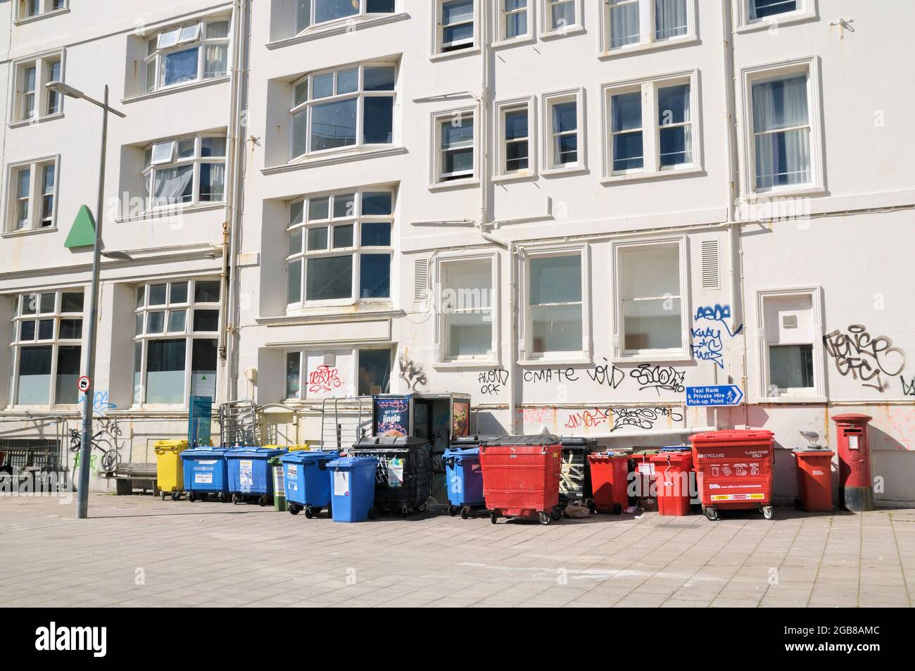 A row of coloured recycling bins and rubbish bins against a white wall partially covered with graffiti scrawl tags, England, UK Stock Photo