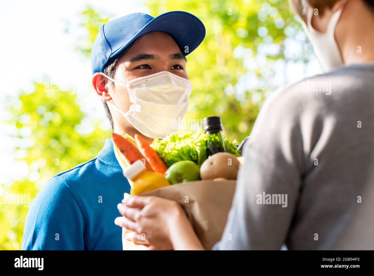 Hygienic Asian deliveryman wearing protective face mask while delivering groceries to customer at home, food delivery in the time of pandemic concept Stock Photo