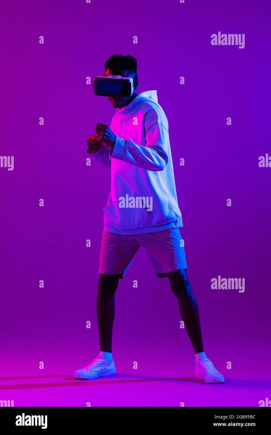 Full length portrait of young African man wearing VR glass headset standing and punching in futuristic dark purple cyberpunk neon light background Stock Photo