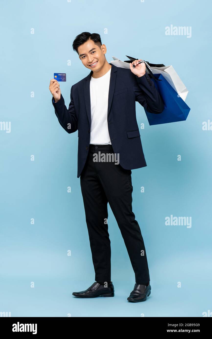 Full length portrait of handsome southeast Asian millenial man holding shopping bags and credit card in isolated studio light blue background for cash Stock Photo