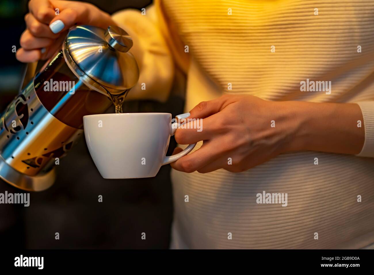 the hand of a pregnant girl holds a white mug, into which she pours tea from a beautiful teapot for brewing in a dark room of the restaurant Stock Photo