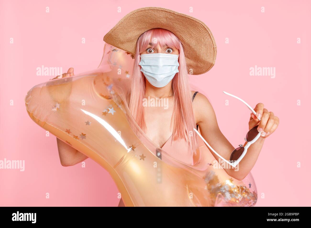 Shocked woman in straw hat and swimsuit with inflatable rubber ring wearing hygienic face mask to prevent contagious coronavirus on resort beach Stock Photo