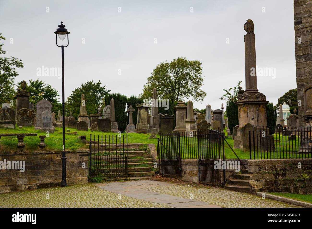 Cementerio de corte gótico con lápidas en forma de piedra y monolitos, en pradera con entrada de barrotes de hierro e iglesia detrás. Stock Photo