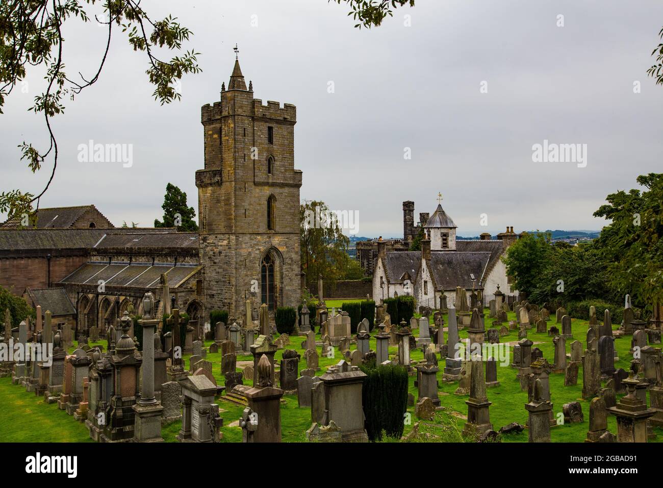 Cementerio de corte gótico con lápidas en forma de piedra y monolitos, en pradera con entrada de barrotes de hierro e iglesia detrás. Stock Photo
