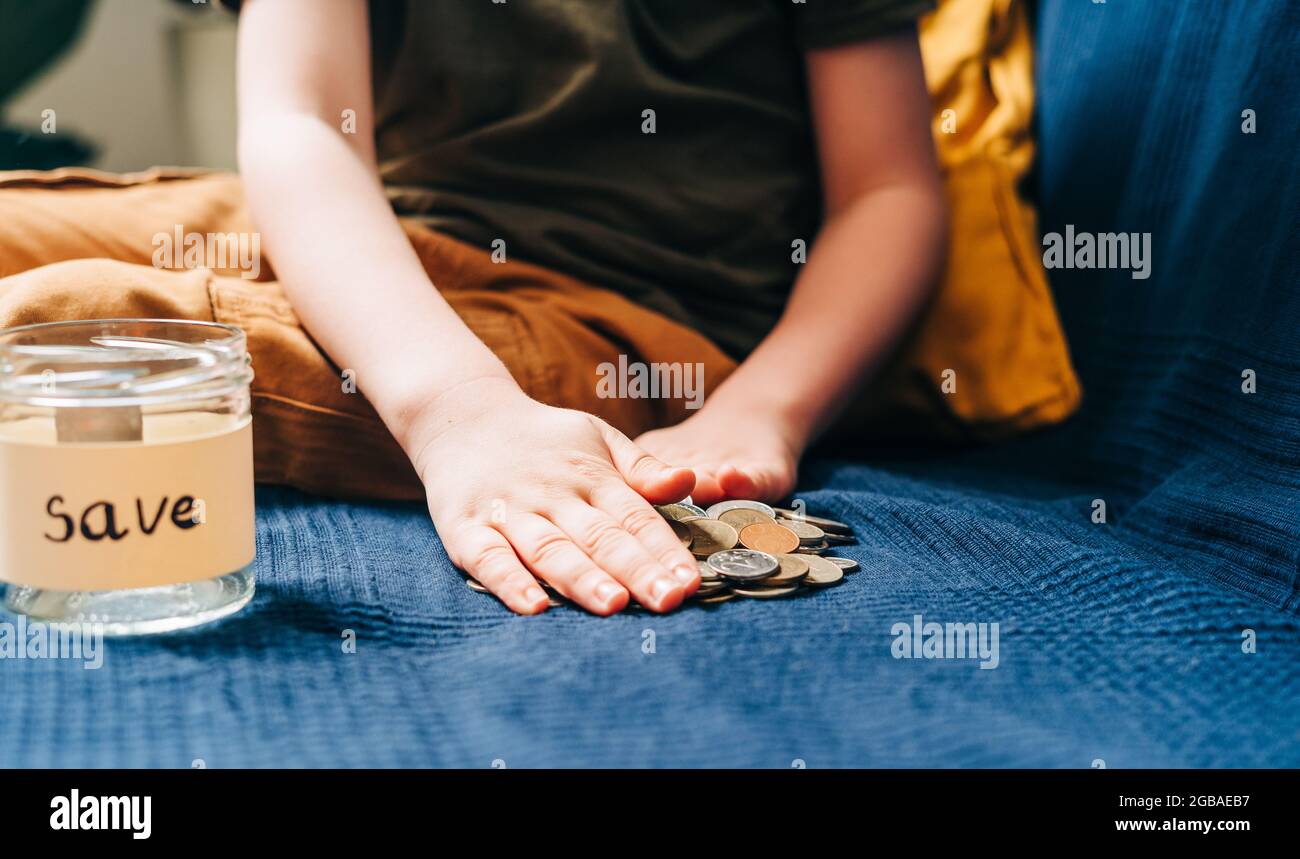 Close up of Little child kid boy hands grabbing and putting stack coins in to glass jar with save label. Donation, saving money, charity, family Stock Photo